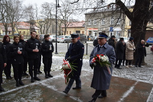 na zdjęciu komendant powiatowy policji w tarnowskich górach i dyrektor aresztu śledczego w trakcie składania wiązanki kwiatów pod pamiątkową tablicą przy kościele świętego piotra i pawła, obok grupa młodzieży