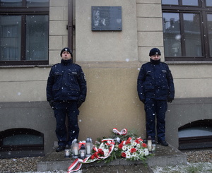 na zdjęciu pamiątkowa tablica na budynku komendy, przy której wartę honorową pełnią policjanci, na ziemi kwiaty i znicze