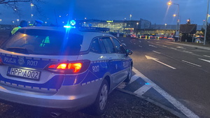 na zdjęciu policyjny radiowóz z włączonymi niebieskimi światłami błyskowymi, w oddali lotnisko w pyrzowicach