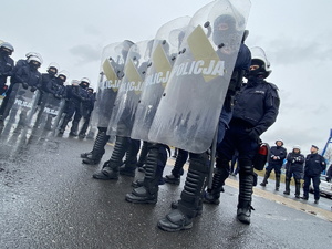 na zdjęciu policjanci z uniesionymi tarczami z napisem policja. stoją w szeregu czteroosobowym. w tle inni policjanci