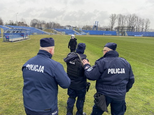 na zdjęciu prowadzący zajęcia policjanci, tyłem do aparatu, wspierają ćwiczącego trzymając dłonie na jego plecach, osoby stają na murawie stadionu, przed ćwiczącymi inny policjant w białym kasku