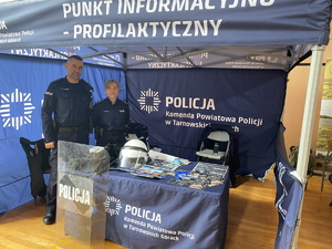 na zdjęciu policjantka profilaktyki i dzielnicowy w namiocie policyjnym, przy stoliku na którym leży policyjny sprzęt i ulotki