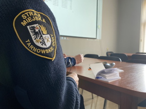 na zdjęciu naszywka straży miejskiej w tarnowskich górach, na blacie notatnik, w tle ekran na którym wyświetlane są dane