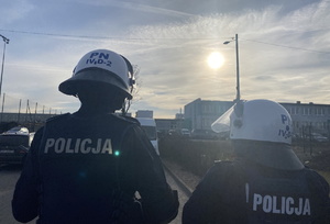 na zdjęciu policjanci w białych kaskach stojący tyłem do obiektywu, przed nimi samochody i budynki