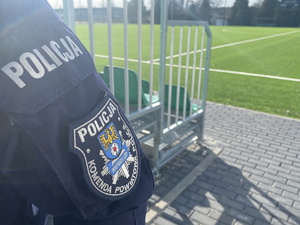 na zdjęciu fragment policyjnego munduru, na którym znajduje się naszywka z napisem Policji Komenda Powiatowa Policji Tarnowskie Góry, w tle murawa boiska