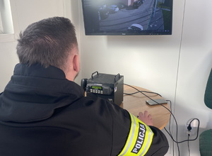 na zdjęciu policjant z opaską na ramieniu z napisem policja, siedzi przy biurku a przed nim monitor, który wyświetla obraz z kamer stadionu