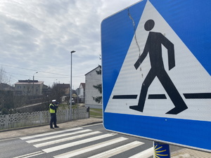 na zdjęciu znak informacyjny przejście dla pieszych, za którym widoczne przejście, obok którego policjant mierzy prędkość