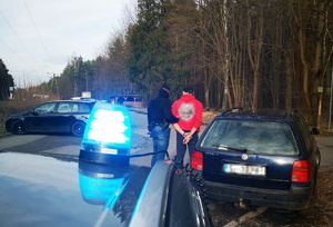 na zdjęciu policjant w ubiorze cywilnym trzyma za rękę zatrzymanego, skutego kajdankami, osoby stoją na parkingu przy pojeździe