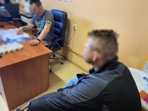 na zdjęciu policjant przy biurku z opaską na napisem policja, obok biurka zatrzymany w ciemnej bluzie