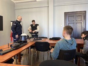 na zdjęciu policjant i jeden z uczniów, który ubiera policyjny sprzęt, obok przy stole inny uczeń