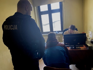 na zdjęciu policjant przy zatrzymanym, który siedzi przy biurku, za biurkiem policjant po cywilu