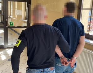 na zdjęciu policjant z opaską z napisem policja prowadzi zatrzymanego korytarzem komendy