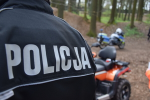 na zdjęciu fragment munduru policjanta z napisem policja, za nim quad i policyjny motocykl