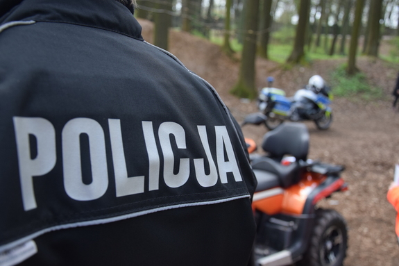 na zdjęciu fragment munduru policjanta z napisem policja, za nim quad i policyjny motocykl