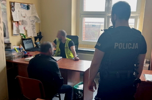 na zdjęciu policjant w mundurze stoi przy zatrzymanym mężczyźnie, który siedzi przy biurku, za którym znajduje się policjant w odblaskowej kamizelce