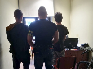 na zdjęciu policjant z opaską z napisem policja na lewym ramieniu stoi pomiędzy dwoma zatrzymanymi poszukiwanymi w pomieszczeniu komendy