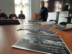 na zdjęciu ulotka informacyjna na stole, za którym stoi policjantka, obok za stołem uczniowie siedzący na krześle