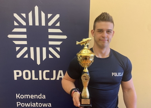 na zdjęciu sierżant Schoppa w mundurze z pucharem w ręku, przy niebieskim banerze z napisem policja