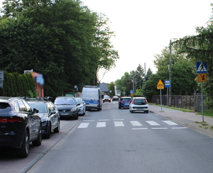 na zdjęciu fragment drogi, na którym widać zaparkowane i jadące pojazdy, pośród nich policyjny radiowóz, w około budynku i drzewa