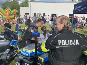 na zdjęciu policjant przy motocyklu, na którym siedzi dziecko, a obok niej rodzic, w oddali inne osoby