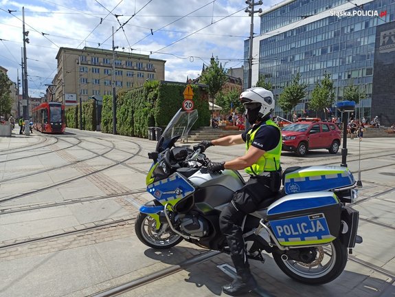na zdjęciu policjant na motocyklu na jezdni i torowisku, za nim tramwaj, samochód i budynki
