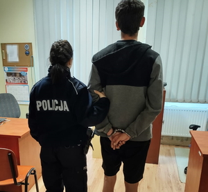 na zdjęciu policjanta trzyma zatrzymanego za ramię w pomieszczeniu komisariatu