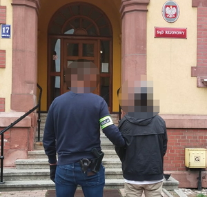 na zdjęciu policjant z opaską na ręku prowadzi zatrzymanego do budynku sądu rejonowego