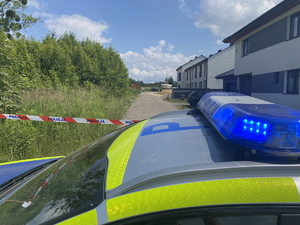 na zdjęciu dach radiowozu i włączone niebieskie sygnały, za nim taśma z napisem policja i budynki oraz samochody