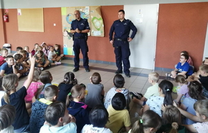 na zdjęciu dzielnicowi stoją przed grupą dzieci, które siedzą na ziemi