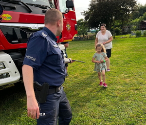 na zdjęciu policjant przy wozie strażackim w trakcie wręczania dziecku kolorowanki