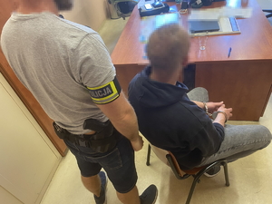 na zdjęciu policjant z opaską na ramieniu, stoi przy zatrzymanym, który siedzi na krześle przy biurku, na rękach ma kajdanki