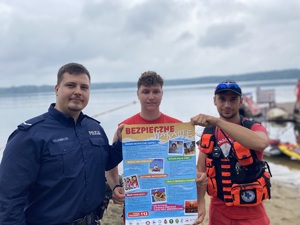 na zdjęciu dzielnicowy z ratownikami trzymają plakat z napisem bezpieczne wakacje, za nimi akwen wodny