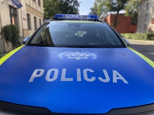 na zdjęciu maska radiowozu z napisem policja, odznaka i hasłem pomagamy i chronimy, w tle rozmazane budynki i drzewa
