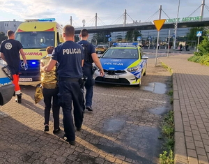 na zdjęciu policjanci prowadza starszą kobietę, okrytą kocem termicznym do karetki, przed nimi idzie ratownik, obok stoi radiowóz policyjny, w tle widoczny terminal lotniska