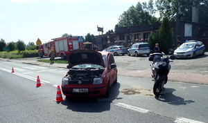 na zdjęciu czerwony fiat na środku jezdni, obok niego uszkodzony motocykl, z tyłu policyjne radiowozy i wóz strażacki, budynki