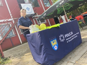 na zdjęciu policjantka za stołem koloru niebieskiego z napisem komenda powiatowa policji w tarnowskich górach, na której leżą gadżety profilaktyczne i balony