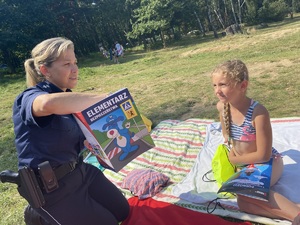 na zdjęciu dziewczyna na kocu, a przed nią policjantka która trzyma w ręku książkę z napisem elementarz bezpieczeństwa