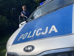 na zdjęciu maska policyjnego radiowozu, a za nią policjant stojący przy drzwiach