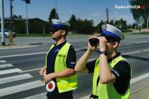 na zdjęciu policjanci na drodze, jeden trzyma w rękach tarcze do zatrzymywania pojazdów, a drugi patrzy przez lornetkę