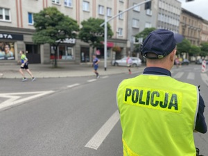 na zdjęciu policjant w odblaskowej kamizelce stoi na jezdni i obserwuje biegaczy, za którymi widać samochody i budynki