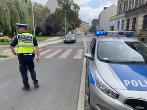 na zdjęciu policjant na jezdni, obok radiowozu, a przed nim czarny samochód osobowy, za którym widoczne są inne pojazdy i budynki