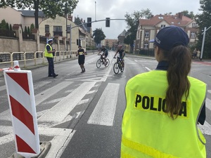 na zdjęciu na pierwszym planie policjantka, która obserwuje biegacza i dwóch rowerzystów na trasie wyścigu, którzy przebiegają obok policjanta stojącego na jezdni