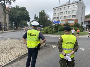 na zdjęciu policjant, obok którego stoi przedstawiciel innej służby mundurowej. mundurowi obserwują teren ronda, przez które przebiegają uczestnicy zawodów
