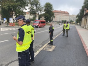 na zdjęciu na pierwszym planie policjanci, obok nich strażak i przedstawiciele innej służby mundurowej. Wszyscy stoją na chodniku i obserwują trasę biegu