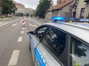 na zdjęciu część policyjnego radiowozu, który na dachu ma włączony neon koloru niebieskiego, stoi zaparkowany na jezdni przy trasie biegu, w tle zabudowania
