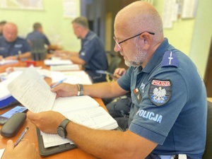 na zdjęciu policjant w trakcie odprawy do służby siedzący przy biurku, w ręku trzyma dokumenty