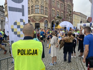 na zdjęciu policjant w odblaskowej kamizelce obserwuje moment startu biegaczy z tarnogórskiego rynku, w tle widoczny tarnogórski ratusz