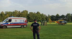 na zdjęciu policjant stojący przodem do zaparkowanej na płycie stadionu karetki pogotowia, obok której stoi śmigłowiec Lotniczego Pogotowia Ratunkowego i strażacy