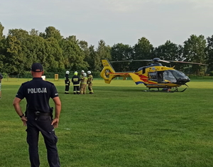 na zdjęciu policjant na płycie stadionu, na którym stoi śmigłowiec Lotniczego Pogotowia Ratunkowego i strażacy