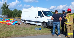 na zdjęciu biały dostawczy samochód, obok niego leżąca na trawie kolorowa plandeka, z drugiej strony stoi policjant, strażak i mężczyzna tyłem w ubiorze cywilnym